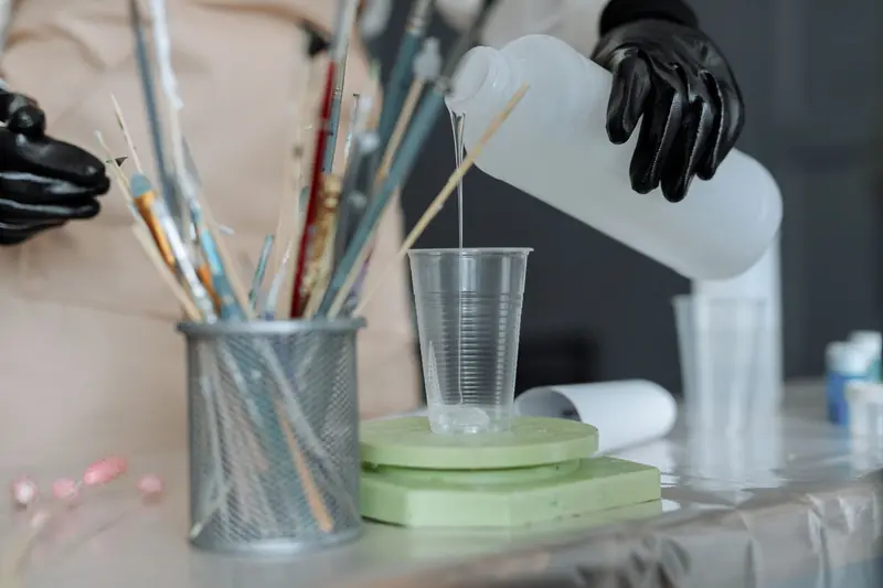 Close-up of resin being poured into a cup amidst art tools and safety gloves