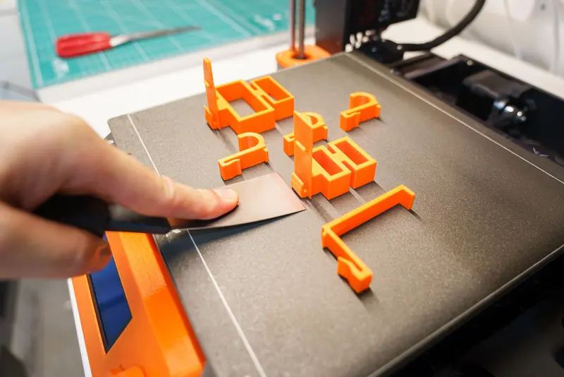 A hand carefully removes an orange 3D printed object from a printer bed using a spatula indoors