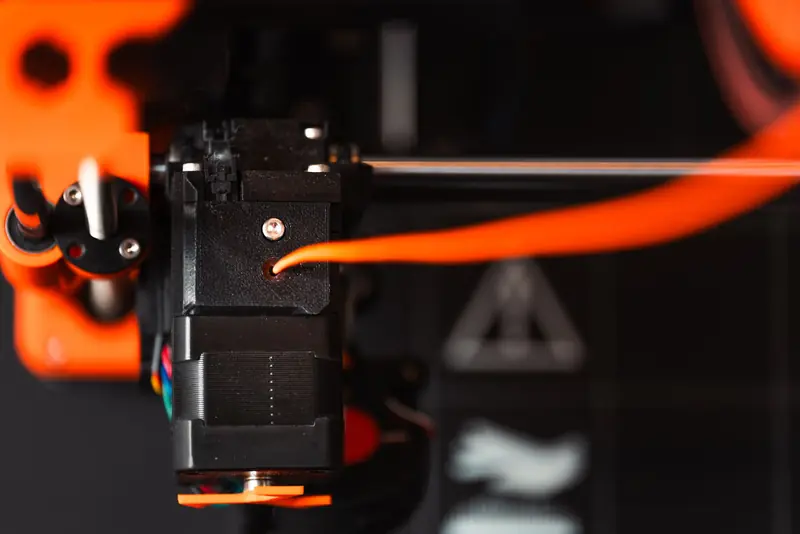 Detailed view of a 3D printer extruder with orange filament in an industrial setting